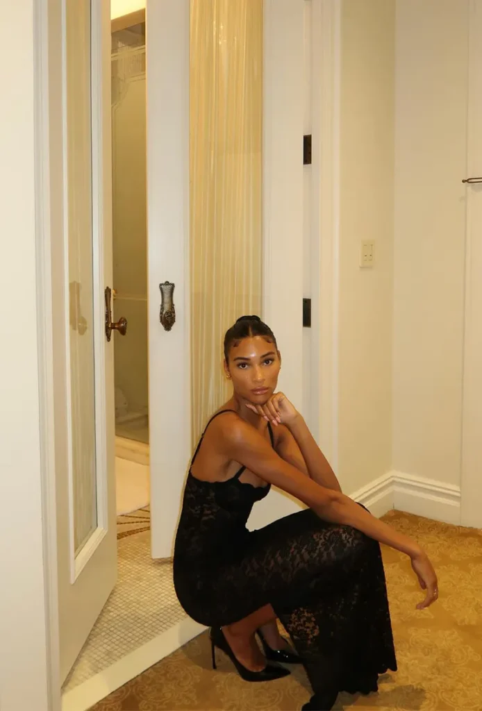 Elegant black lace evening dress styled with pointed heels, worn by a woman seated in a softly lit interior setting.