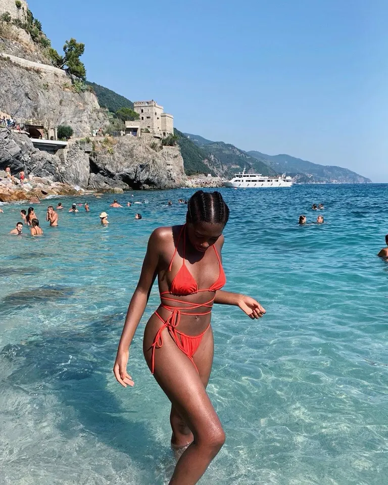 Woman in red bikini walking in clear turquoise water at a rocky coastal beach with cliffs and yacht in background