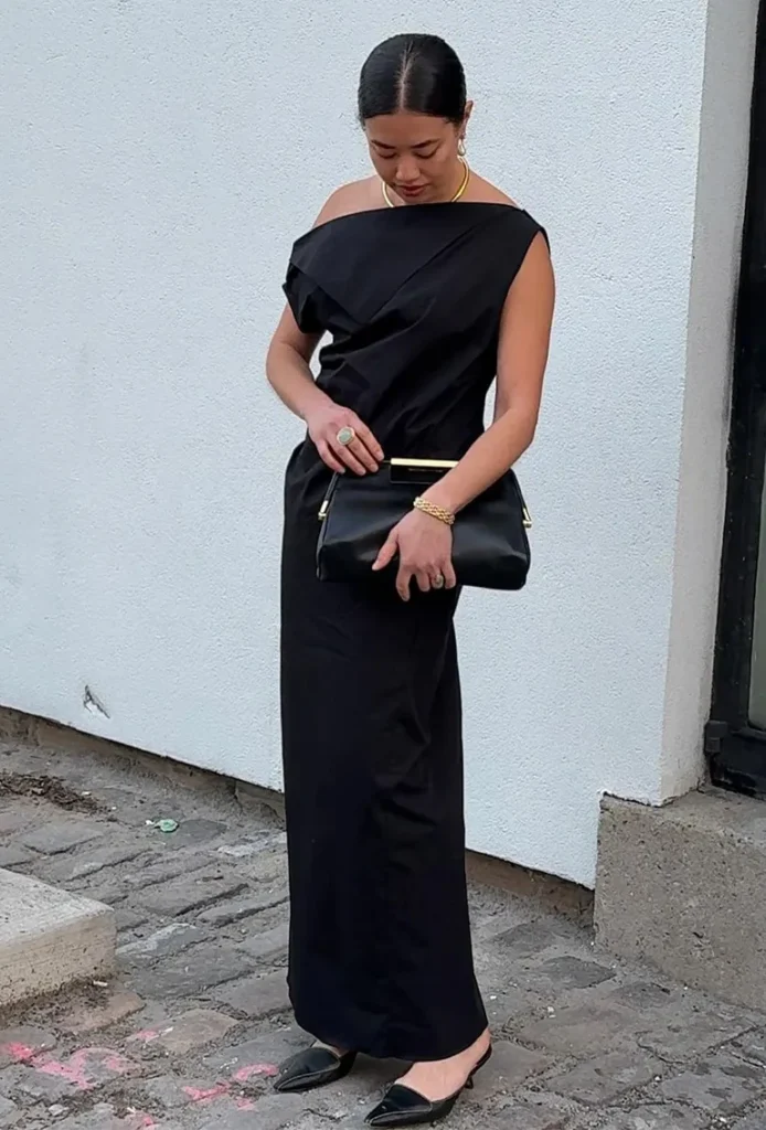 Chic all-black evening outfit featuring a one-shoulder maxi dress, gold accessories, and a structured black clutch.