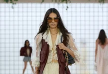 We’re Calling It Now—This Pretty Top Trend Just Might Replace Your White Tee This Season Model walking the runway wearing a cream ruffled mini dress layered with a burgundy leather vest, oversized sunglasses, lace-up heels, and carrying a small shoulder bag.