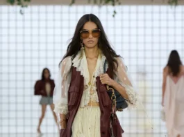 We’re Calling It Now—This Pretty Top Trend Just Might Replace Your White Tee This Season Model walking the runway wearing a cream ruffled mini dress layered with a burgundy leather vest, oversized sunglasses, lace-up heels, and carrying a small shoulder bag.