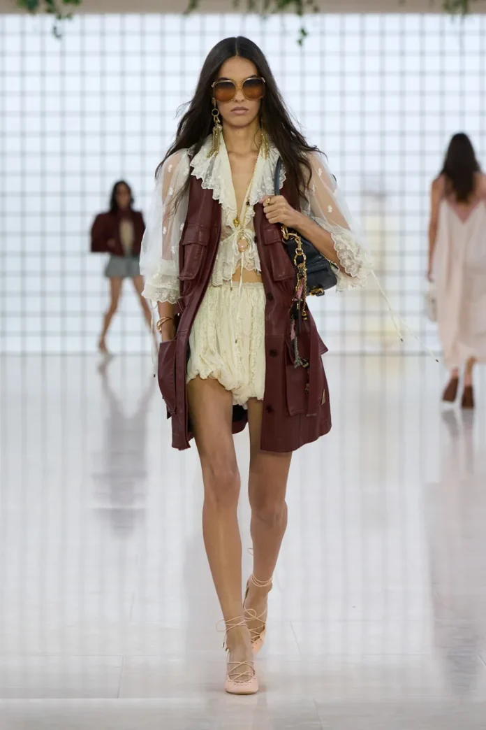 featur Model walking the runway wearing a cream ruffled mini dress layered with a burgundy leather vest, oversized sunglasses, lace-up heels, and carrying a small shoulder bag.