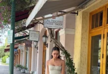 The Elegant, Lingerie-Inspired Dress Trend I’ve Seen All Over Europe This Summer Woman wearing a sage green midi dress with a fitted bodice and flared skirt, styled with strappy heels, walking along a charming outdoor shopping street with boutique storefronts.