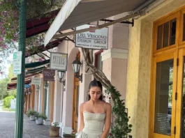 The Elegant, Lingerie-Inspired Dress Trend I’ve Seen All Over Europe This Summer Woman wearing a sage green midi dress with a fitted bodice and flared skirt, styled with strappy heels, walking along a charming outdoor shopping street with boutique storefronts.