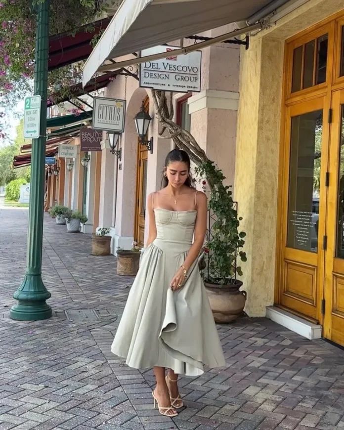 featur img Woman wearing a sage green midi dress with a fitted bodice and flared skirt, styled with strappy heels, walking along a charming outdoor shopping street with boutique storefronts.