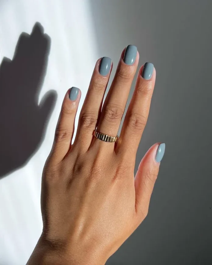 Soft grey-blue glossy manicure with gold ring in natural sunlight