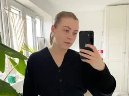 Woman taking a mirror selfie wearing a black button-front cardigan with balloon sleeves and high-waisted black jeans in a bright bedroom with plants and white decor.