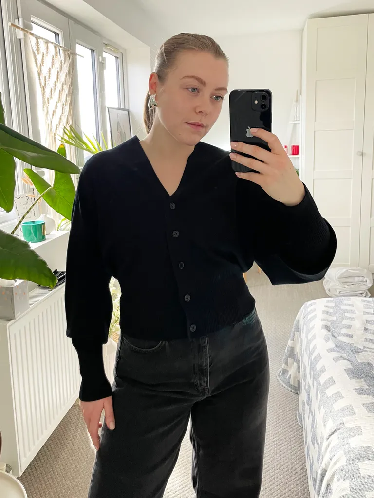 Woman taking a mirror selfie wearing a black button-front cardigan with balloon sleeves and high-waisted black jeans in a bright bedroom with plants and white decor.