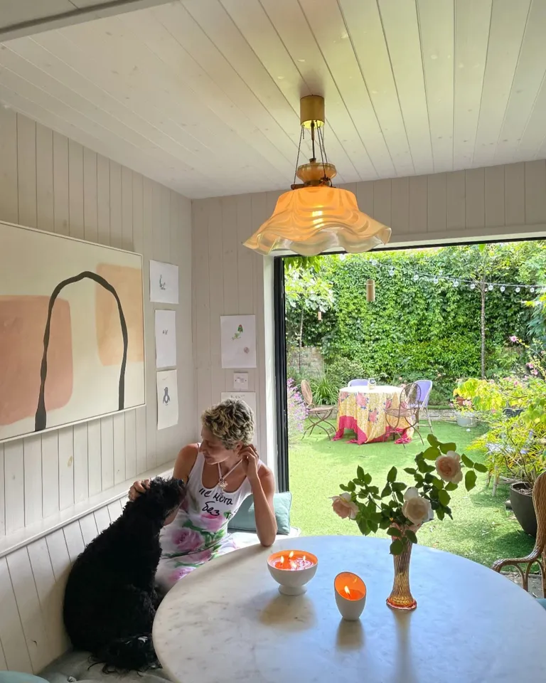 A Person petting black dog at a candle-lit table with flowers, beside an open doorway overlooking a green garden patio.